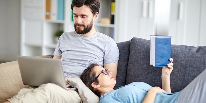 couple se relaxant sur un canapé homme devant un ordinateur pendant que la femme lit un livre