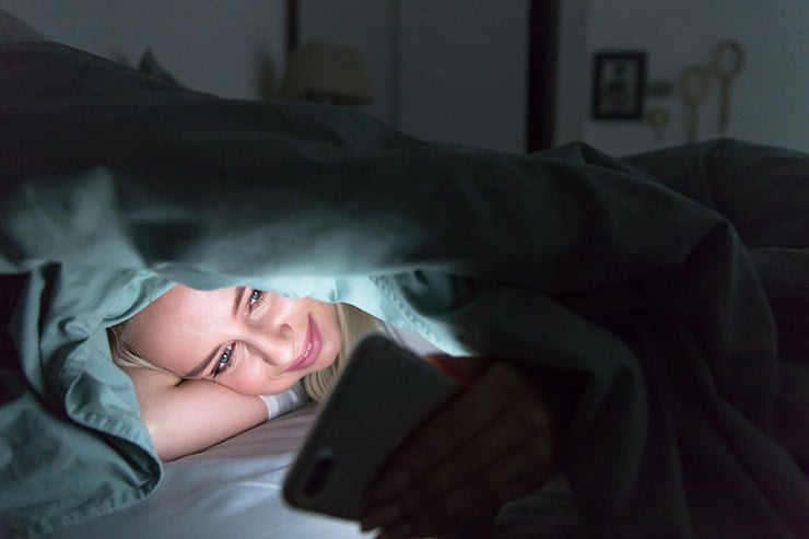 mulher relaxada deitada na cama, debaixo do cobertor, usando seu telefone