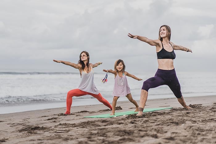 två kvinnor och ett barn som utövar yoga på stranden där det gör yoga warrior 2 posen