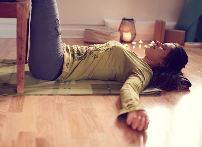 Frau, die eine restaurative Yogapose ausführt, liegt auf dem Boden mit den Beinen auf einem Stuhl.